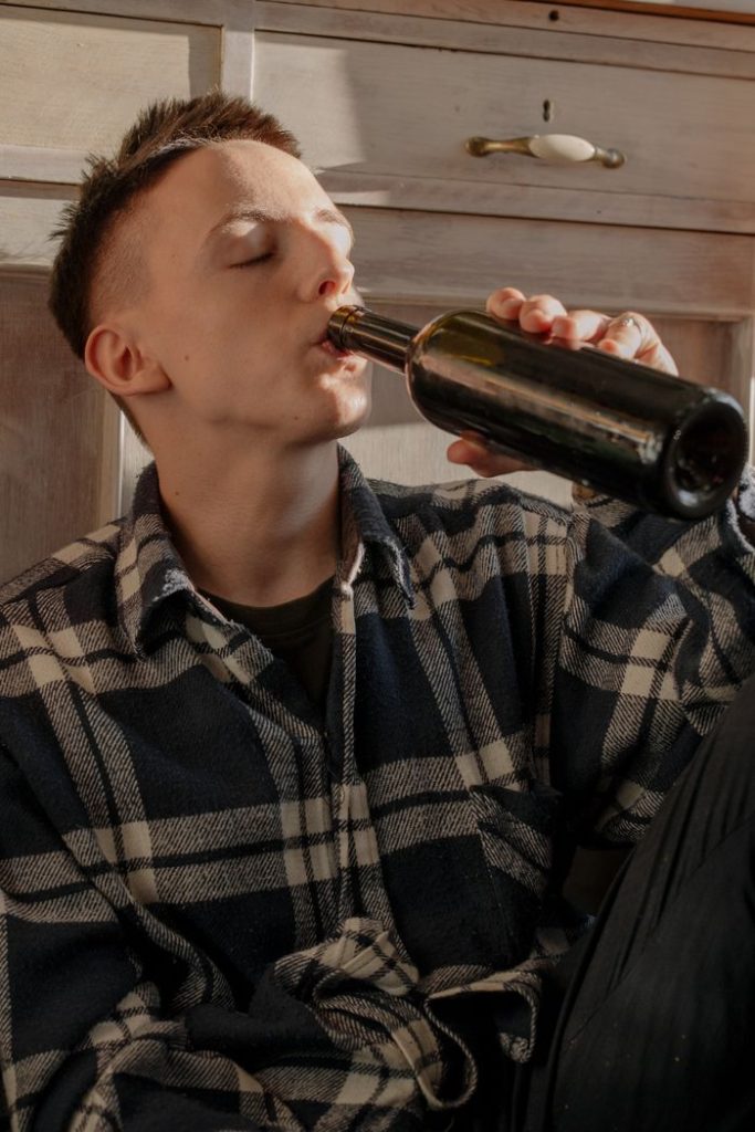 A man in a checkered shirt drinks from a bottle indoors, sitting on the floor.